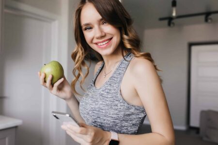 slim gorgeous girl with wavy hair posing wih phone and fruit indoor portrait of romantic female model eating green apple in morning scaled1